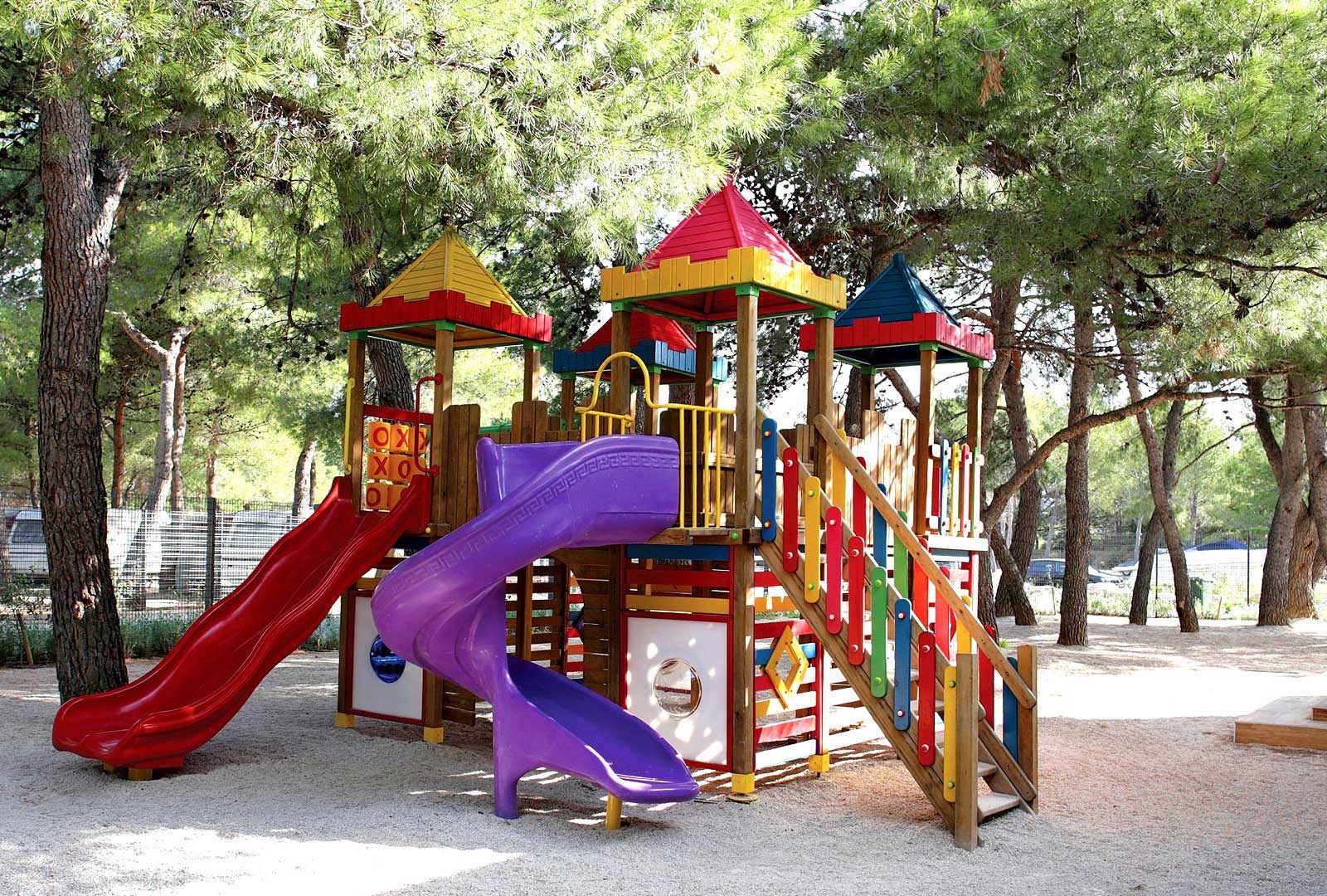 Children's playground with two slides in range of colors standing in the pebbles ground with a  few pine trees surrounding it.